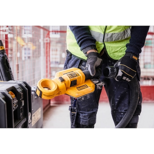 A person wearing a safety vest and gloves is using a DEWALT DWH050 dust extractor attachment at a construction site. The yellow tool is connected to a hose and positioned near a DEWALT storage case.