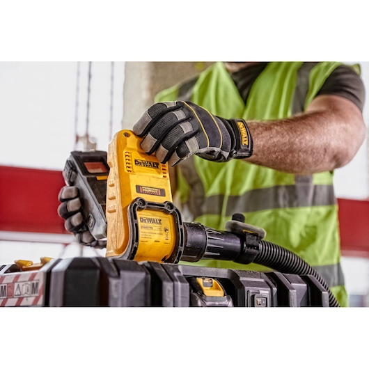 A person wearing work gloves and a safety vest is holding and operating a DEWALT DWH079D dust extraction unit, connecting it