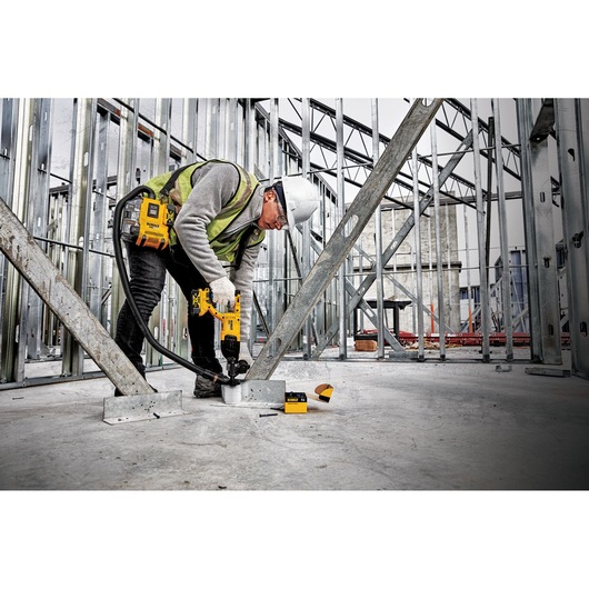 A construction worker using a DEWALT portable dust extractor with a drill inside a building under construction, surrounded