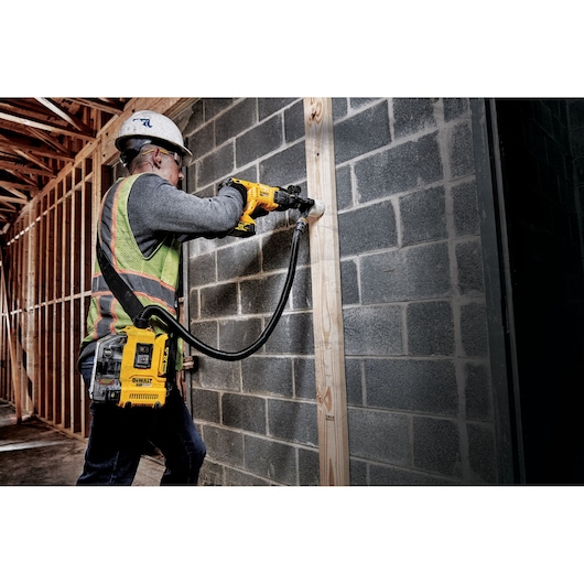 A worker wearing a safety helmet and high-visibility vest is using a DEWALT DWH161D1 dust extractor attached to a cordless