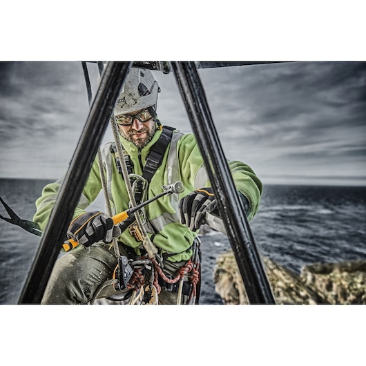 A worker in safety gear and a helmet is suspended on ropes over a coastal area, holding a DEWALT tool. The face is blurred