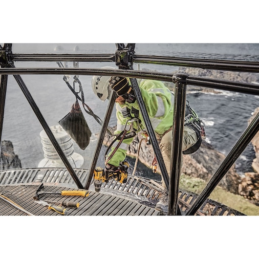 A worker wearing safety gear and a helmet is using a DEWALT drill on the metal framework of a structure located above rocky