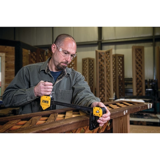 A person using a DEWALT DWHT0-83186 clamp to secure wooden boards in a workshop. The clamp is yellow and black, and the