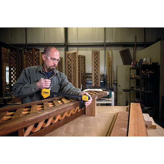 A person in a workshop uses a DEWALT DWHT0-83186 clamp to secure a wooden lattice structure on a workbench. Other wooden