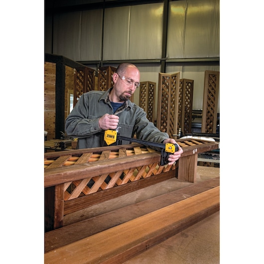 A person uses a DEWALT bar clamp (model DWHT0-83186) to secure a wooden lattice structure on a workbench in a workshop. The