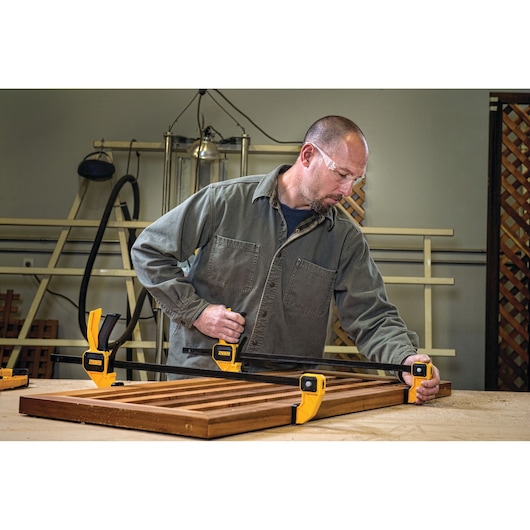 A person working in a workshop using DEWALT clamps (DWHT0-83194) to secure wooden boards on a workbench. The DEWALT clamps