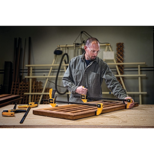 A person working in a workshop using DEWALT bar clamps to secure a wooden frame on a workbench. Several yellow and black