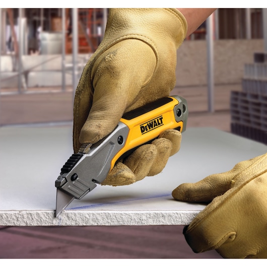 A close-up of a person wearing yellow protective gloves using a DEWALT utility knife to cut a piece of drywall or sheet