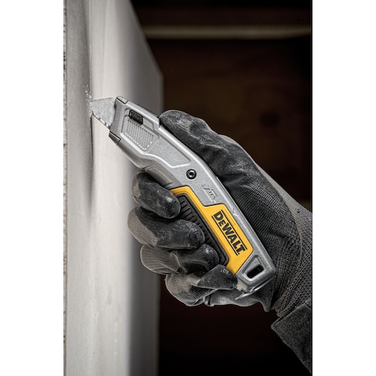 A person wearing black gloves is using a DEWALT utility knife to cut into a sheet of building material.