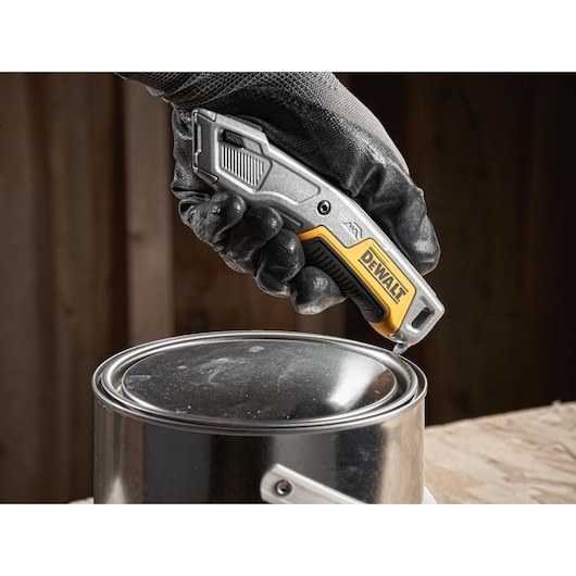 A gloved hand is using a DEWALT utility knife to open the lid of a metal paint can on a wooden surface.
