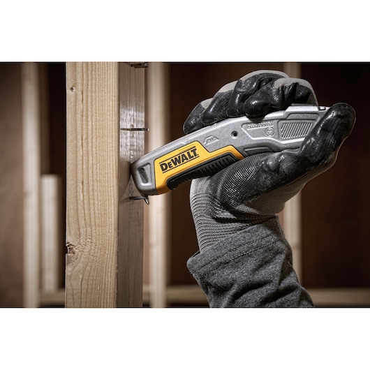 A gloved hand using a DEWALT utility knife (model DWHT10054-0) to cut or score wood in a construction setting.