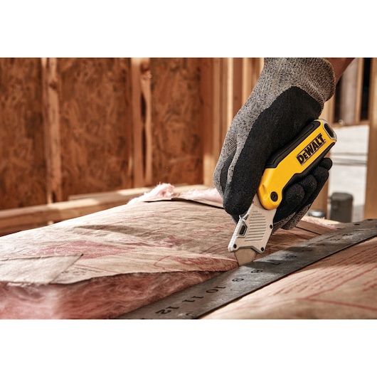 A person wearing work gloves is using a DEWALT utility knife to cut insulation material along a metal ruler inside a