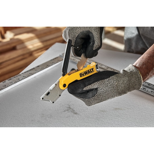 Gloved hands using a DEWALT DWHT10429-0 utility knife to cut material on a work surface.