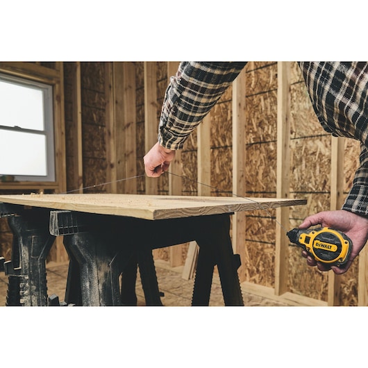 A person using a DEWALT tape measure (model DWHT48333BC) to measure a piece of wood supported by sawhorses in a workshop under construction.