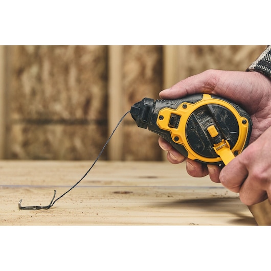 A hand holding a DEWALT chalk line reel, pulling out the string over a wooden surface. The chalk line tool is black and yellow in color, used for marking straight lines on construction surfaces.