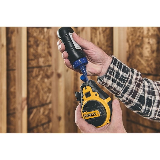 A person wearing a plaid shirt is pouring blue chalk powder from a bottle into a DEWALT chalk line tool against a background of wooden walls.