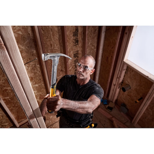 A person using a DEWALT hammer (model DWHT51135-0) to drive a nail into a wooden frame inside a partially constructed room. The hammer has a yellow and metal handle.
