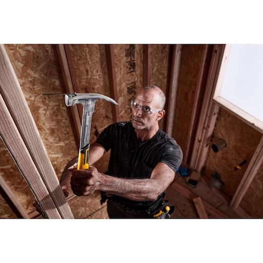 A person using a DEWALT hammer (DWHT51135-0) to work on a wooden frame inside a construction site. The hammer has a yellow and black handle, and the person is wearing a tool belt.