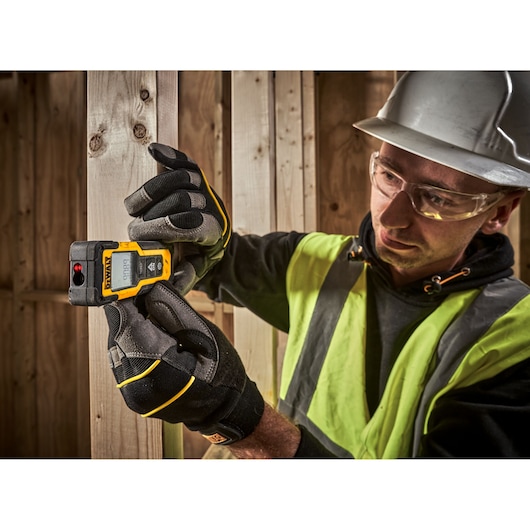 A person wearing safety gloves and a high-visibility vest is using a DEWALT DWHT77100 laser distance measurer to take