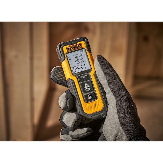 A hand wearing a work glove is holding a DEWALT DWHT77100 laser distance measurer. The device is yellow and black