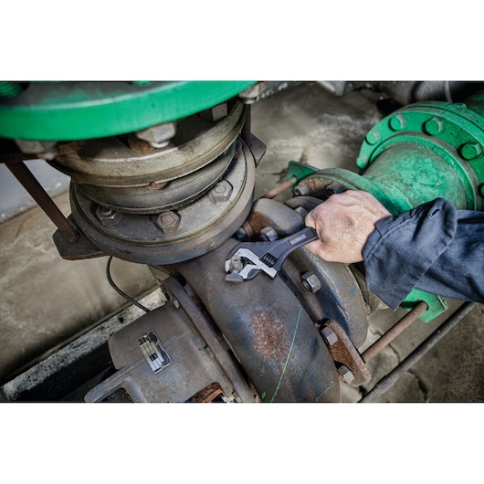 A person uses a DEWALT adjustable wrench to tighten a bolt on a large industrial pipe with metal fittings and flanges.
