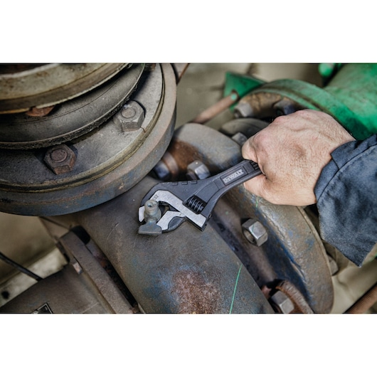A DEWALT adjustable wrench is being used to tighten or loosen a bolt on industrial piping. A hand is gripping the wrench
