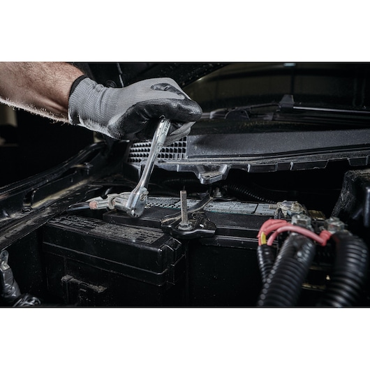 A gloved hand uses a ratchet wrench to tighten or loosen a bolt on a car battery under the hood.
