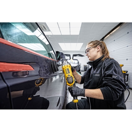 Person using a yellow DEWALT DWP849X polisher on the side of a dark vehicle in an indoor garage setting. The person is wearing black gloves and a black jacket.