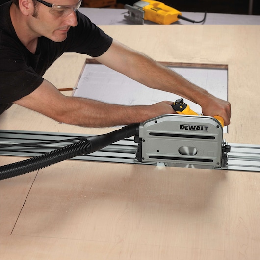 A person wearing safety glasses operates a DEWALT track saw on a wooden surface. The saw is connected to a vacuum hose for dust collection, and there is a rectangular cutout in the wood.