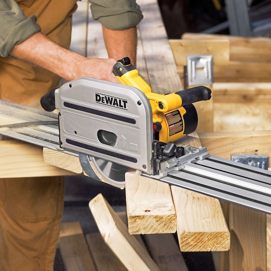 A person using a DEWALT track saw (model DWS520K) to make precise cuts on wooden boards. The saw is positioned on a metal guide rail for accuracy. The workspace includes several pieces of lumber and woodworking materials.