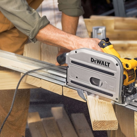 A person is using a DEWALT DWS520K track saw to cut a piece of wood. The saw is positioned on a guide rail for precise cutting.