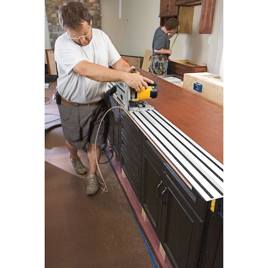 A person is using a DEWALT track saw (DWS520K) to cut a large wooden countertop in a kitchen setting. Another person in the background is measuring an area near the cabinets. The DEWALT saw is positioned on a guide rail for accurate cutting.