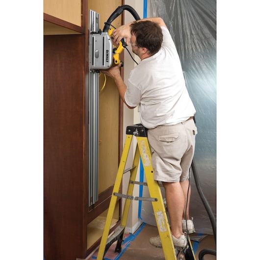 Person using a DEWALT DWS520K track saw on vertical wood panel while standing on a yellow step ladder indoors.