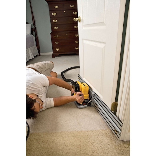 A person using a DEWALT track saw with a guide rail to cut the bottom of an interior door. The scene is set in a carpeted bedroom with furniture visible in the background.