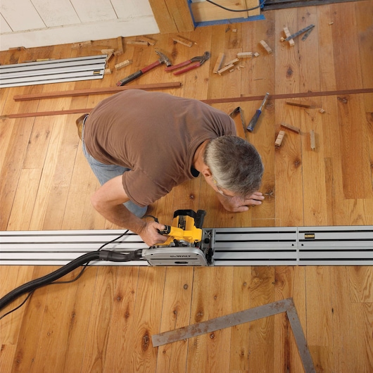 A person using a DEWALT track saw (model DWS520K) on a wooden floor to make a straight cut, with various hand tools and wood pieces scattered around.