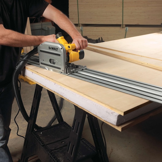 A person using a DEWALT track saw (model DWS520K) to cut a large sheet of wood on a worktable. The saw is guided by a metal track for precise straight cuts.