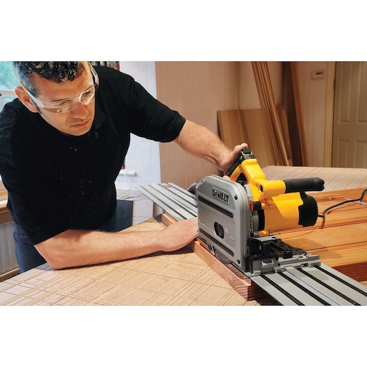 A person using a DEWALT DWS520K track saw to cut wood on a workbench. The saw is positioned on a metal guide rail for precise cutting. The workspace includes wooden boards and tools.
