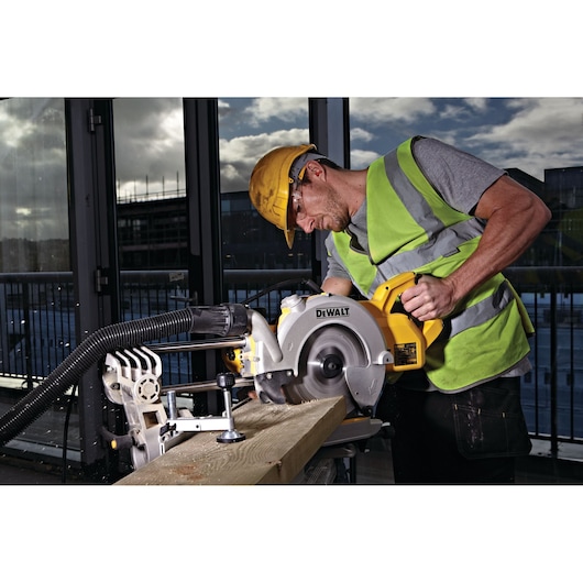 Worker using DEWALT DWS778 saw to cut wood on a construction site, wearing safety gear and hi-vis vest.