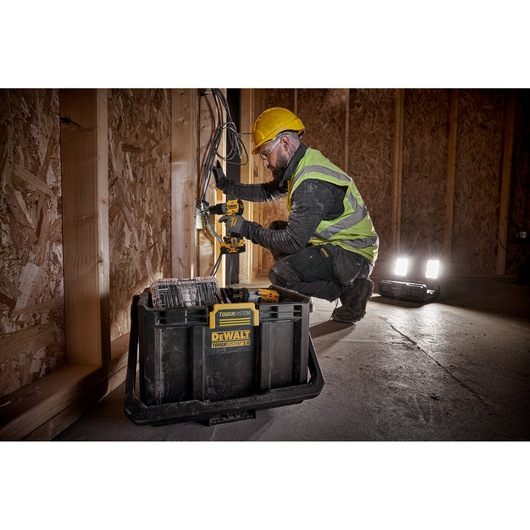 A person wearing a yellow hard hat and a high-visibility vest is using a DEWALT power tool to work on wiring inside a wooden construction site. A DEWALT TOUGHSYSTEM 2.0 toolbox is prominently placed in the foreground, and portable work lights are illuminating the area.