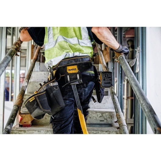 A construction worker wearing a high-visibility vest climbs stairs at a worksite, carrying a DEWALT tool belt equipped with