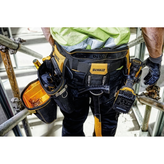 Close-up view of a person wearing a DEWALT tool belt, loaded with various tools, fasteners, and a cordless power tool. The