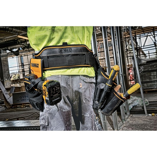 A close-up view of a person wearing a DEWALT tool belt loaded with various tools, standing on a construction site. The tool belt features multiple compartments and pockets holding screwdrivers, a battery pack, and other hand tools. The background shows scaffolding and construction materials.