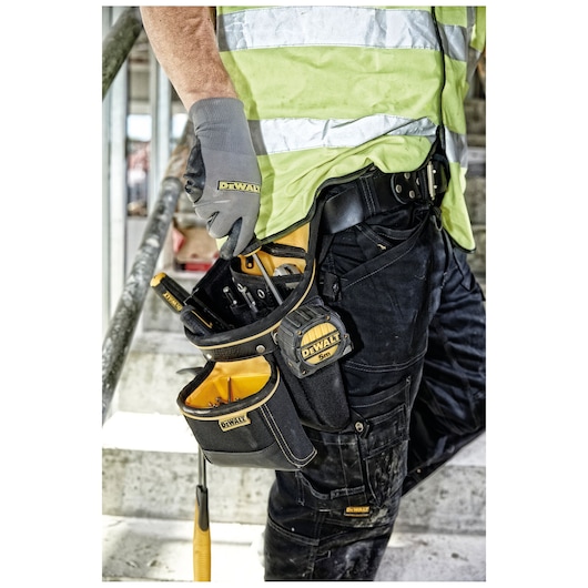 Close-up of a worker wearing a DEWALT tool belt and gloves, equipped with various tools and a measuring tape, standing on a