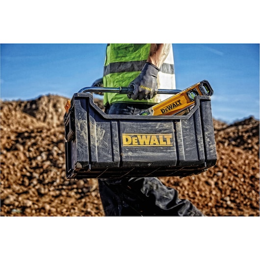 A person wearing gloves and a high-visibility vest carries a black DEWALT toolbox with a level tool inside, on a construction site with dirt mounds in the background.