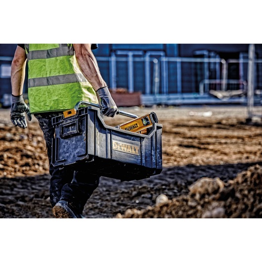 A person wearing a safety vest and gloves is carrying a black DEWALT toolbox with a handle and DEWALT tools inside, walking across a construction site.