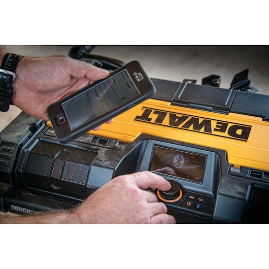 A DEWALT ToughSystem portable radio and charger is being operated by someone adjusting the control knob and holding a