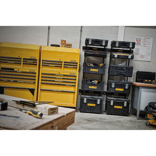 A workshop scene featuring yellow DEWALT tool storage cabinets and several stacked black DEWALT toolboxes against a wall. A workbench with tools is visible in the foreground.