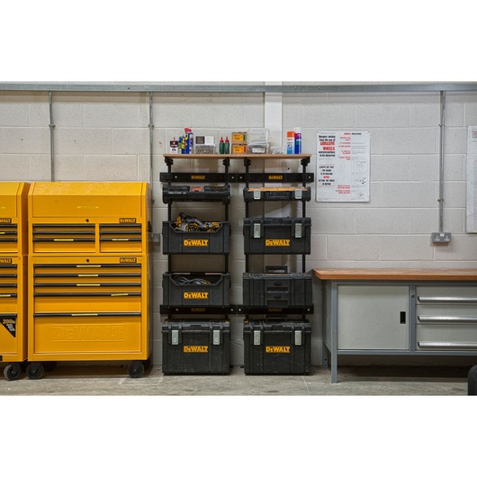 A workshop scene showing DEWALT storage solutions. On the left, there are large yellow tool cabinets with multiple drawers. In the center, black DEWALT toolboxes are stacked on a wall-mounted shelving system with tools and supplies on the shelves. On the right, there is a workbench with metal drawers. The workshop has grey brick walls with safety posters and power outlets.