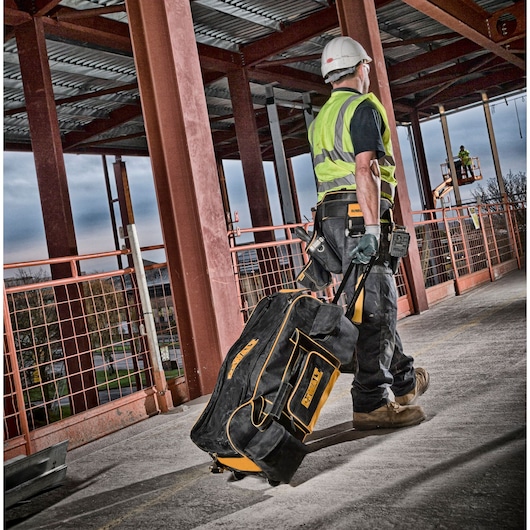 A construction worker wearing a helmet and high-visibility vest pulls a DEWALT DWST1-79210 rolling tool bag across a