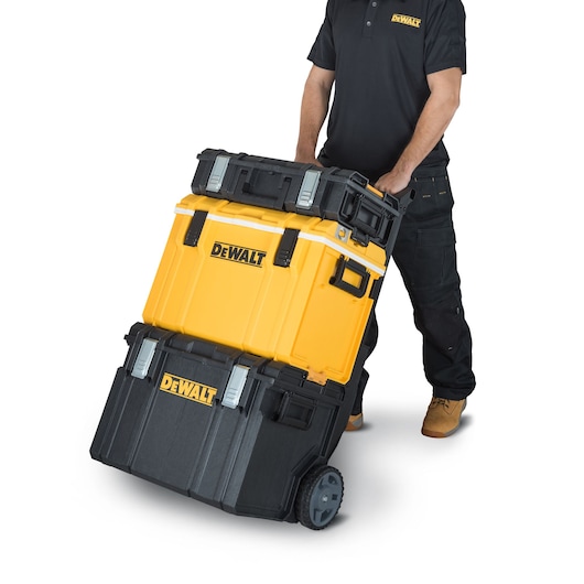 A person wearing black work clothing and tan boots is pulling a DEWALT portable tool storage system with three stacked compartments on wheels.